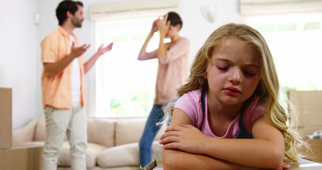 Image depicts a young girl feeling sad due to her parents' argument in their living room. Used in articles or campaigns about family dynamics, childhood, emotional health, therapy, parenting challenges.