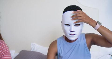 Young african american person adjusting led facial mask while wearing smartwatch in bedroom
