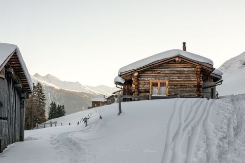 Snow-covered log cabin nesting on alpine slope with soft morning light and mountain peaks