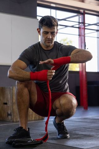 Mid-adult man wrapping red hand wraps before boxing workout in gym, preparing for training