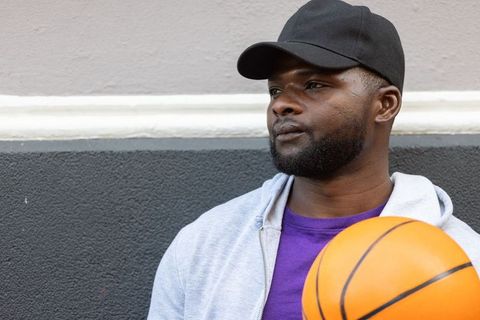 Urban african man holding basketball, leaning on two-tone wall wearing cap and hoodie