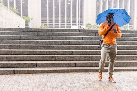 Walking on wet urban steps holding blue umbrella and checking smartphone, orange sweater