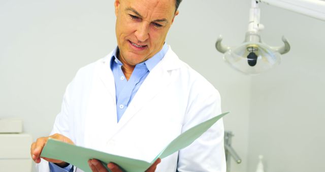 Male doctor in white coat holding and reviewing patient medical records in bright, well-lit clinical setting. Suitable for illustrating healthcare, medical consultations, clinical procedures, and professionalism in medical industry-related content. Useful for medical websites, healthcare brochures, and educational materials.