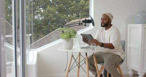 Man hosting home podcast in bright minimal studio using tablet and microphone