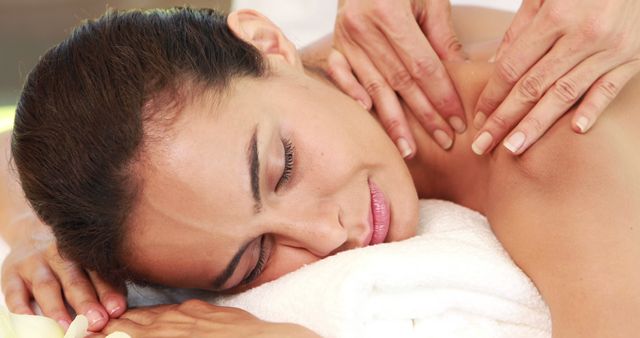 A woman lays peacefully on a white towel while receiving a professional massage. Her eyes are closed, and she appears to be experiencing deep relaxation as the therapist's hands work on her back. This image can be used for advertising spa services, wellness and health publications, or any content related to stress relief and pampering.