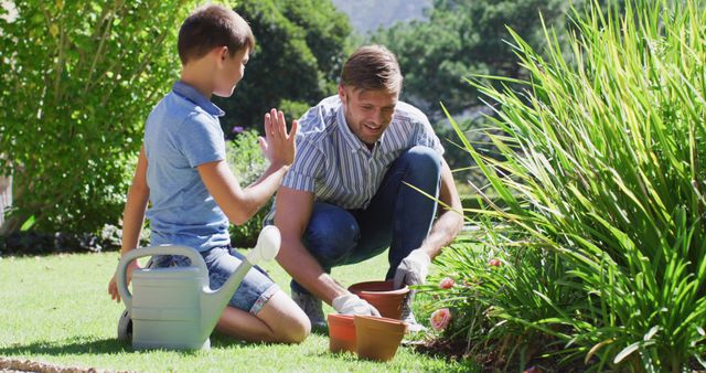 Father and son enjoying bonding time while gardening in sunny backyard. Father planting while son holds watering can. Ideal for content related to family activities, outdoor hobbies, gardening tips, or parenting.