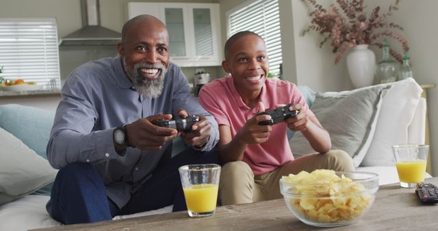 Father and son enjoy quality time playing video games in their cozy living room. Their joyful expressions and the casual ambiance make this ideal for promoting family activities, gaming consoles, or leisure time advertisements. Perfect for depicting family bonding and joyful moments. The presence of snacks and drinks adds a relatable, homey touch.