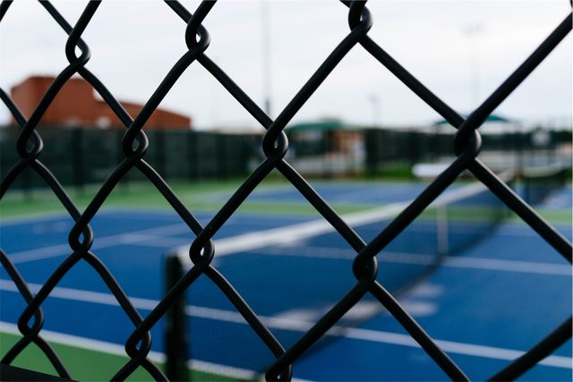 This image shows an empty tennis court as seen through the grid of a chain-link fence. The background of the shot includes blue and green court surfaces and sports equipment, with the fence being the main focus of the image. The image can be useful for illustrating themes such as sports facilities, recreational activities, or urban sports environments. It is suitable for use in articles related to sports events, fitness, or community recreational spaces.