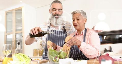 Preparing mature couple cooking together, tossing greens and drizzling olive oil