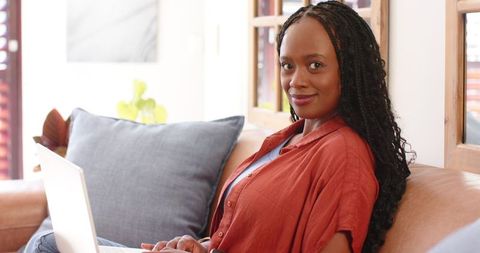 African american woman working remotely on laptop at home on sofa with natural light