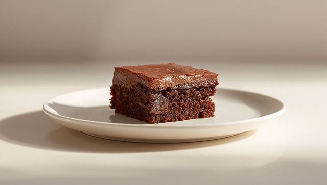Fudgy chocolate cake square with glossy ganache sitting on white ceramic plate