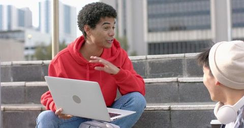 Urban campus students collaborating on laptop outdoors, casual discussion on steps