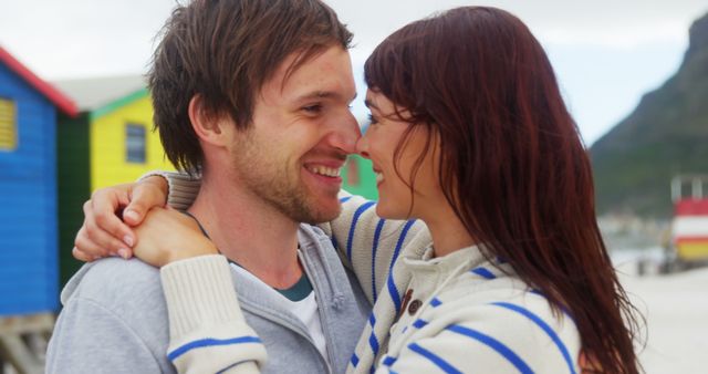 A young Caucasian couple shares a close embrace on a beach with colorful beach huts in the background. Their affectionate gaze suggests a romantic connection amidst a scenic coastal setting.