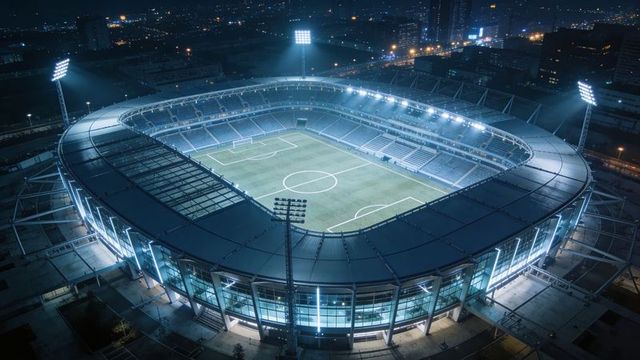 Illuminated modern soccer stadium shining over urban skyline at night, aerial perspective