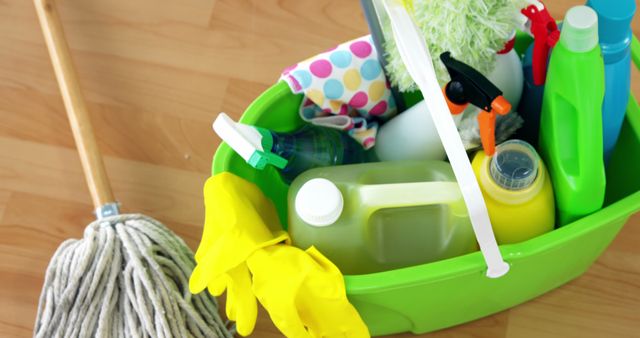 Various cleaning supplies in a green bucket displayed on a wooden floor. Items include a mop, yellow rubber gloves, detergent bottles, sprayers, and a colorful cloth, representing comprehensive house cleaning tools. Ideal for illustrating cleaning services, tutorials on house cleaning, or advertisements for cleaning products.