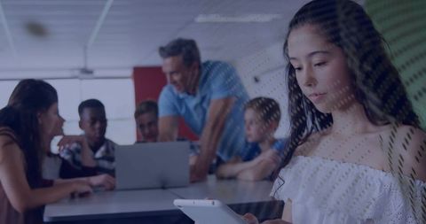 Teen using tablet in collaborative classroom while teacher and peers working on laptop