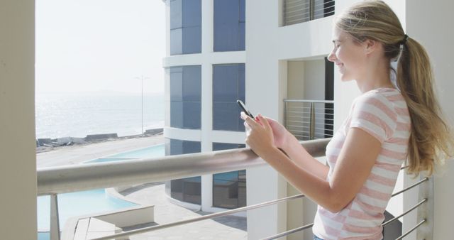 Young woman standing on balcony of modern beachside apartment, texting on smartphone while enjoying sunrise over ocean. Suitable for relaxation, technology, vacation, coastal living themes, or advertisements emphasizing leisure lifestyle and connectivity.