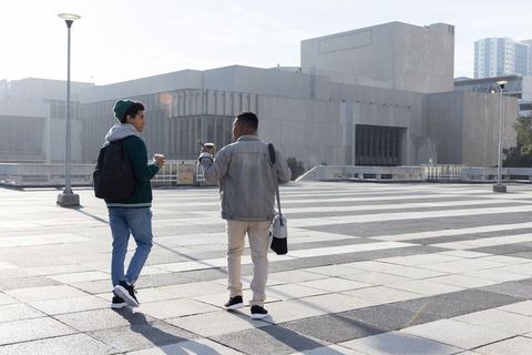 Two young men walking and talking across urban plaza with coffee, backpacks, smartphone
