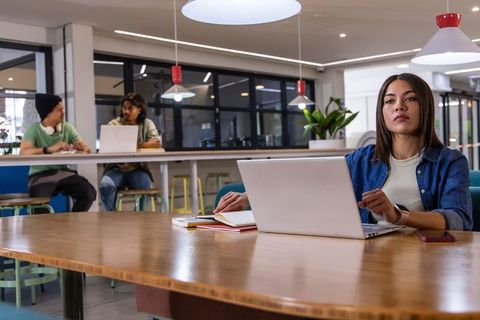 Focused woman working on laptop at coworking communal table with notebooks phone
