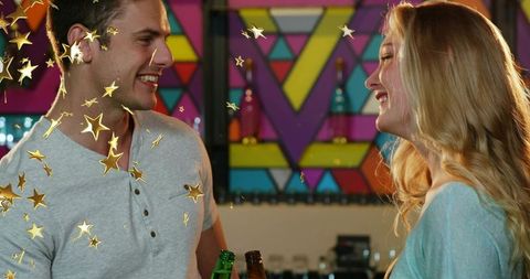 Couple clinking beer bottles at colorful bar with gold star confetti and mural