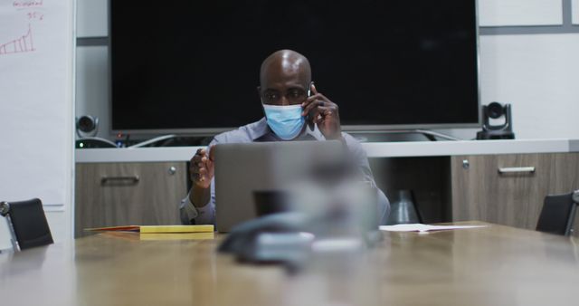 African american businessman in face mask talking on smartphone using laptop in conference room. business professional working in modern office during covid 19 coronavirus pandemic.