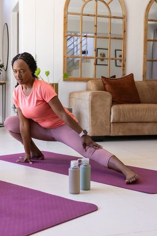Woman stretching side lunge on yoga mat in sunlit living room in coral top and mauve leggings