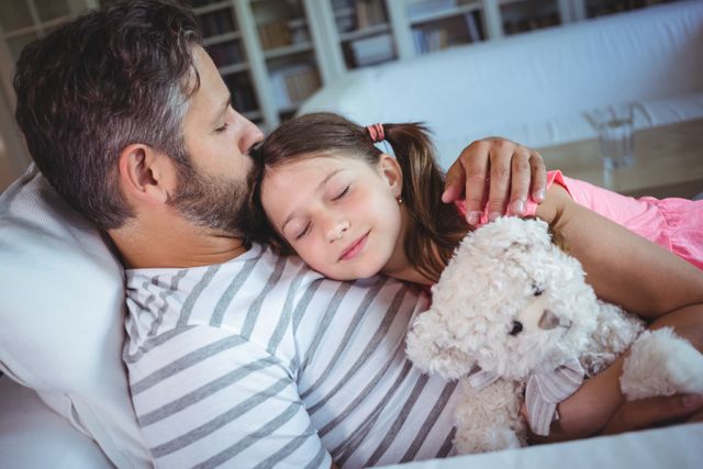 Father and daughter sharing a tender moment at home, with the daughter sleeping in her father's arms while holding a teddy bear. Ideal for use in family-oriented advertisements, parenting blogs, and articles about father-daughter relationships and family bonding.