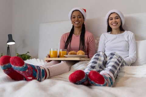 Cozy holiday morning in bed with diverse women wearing santa hats holding breakfast tray