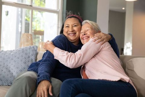 Mature women hugging and smiling on cozy sofa near sunlit window, warm companionship