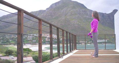Barefoot woman holding purple dumbbells on wooden deck facing bay and mountain view