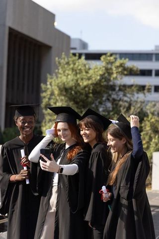 Diverse female graduates taking campus selfie in caps and gowns celebrating together