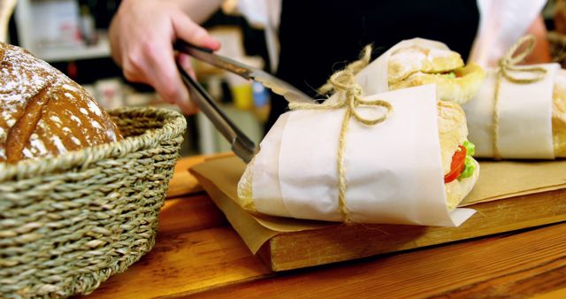 Person using tongs to place prepared sandwiches wrapped in paper and tied with string on wooden surface. Nearby, basket filled with freshly baked bread. Suitable for illustrating homemade food preparation, cafeteria settings, lunchtime concepts, or bakery advertisements.