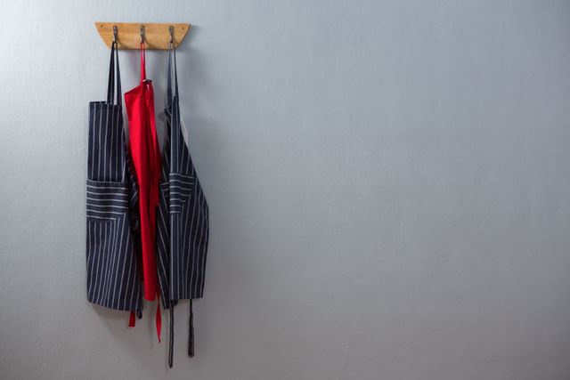 Various aprons hanging on a wooden hook against a plain wall. Ideal for illustrating kitchen organization, culinary workspaces, or home decor. Suitable for articles on cooking, chef uniforms, or interior design tips.