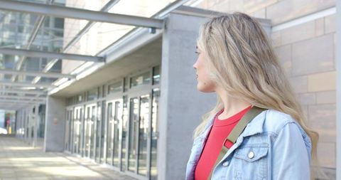 Young woman standing by modern glass building wearing denim jacket and red top