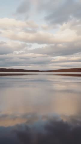 Drifting clouds mirroring on calm lake at sunrise over low hills, vertical video