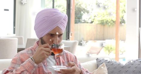 Turbaned man sipping tea in sunlit living room with sliding doors and backyard view