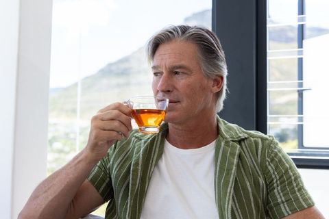 Mature man sitting in sunroom sipping tea, contemplative moment overlooking hillside