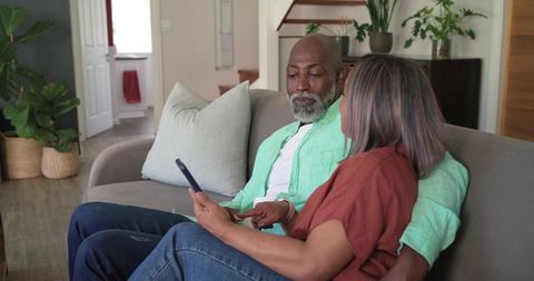 Mature couple sharing tablet on cozy gray sofa in modern living room with plants