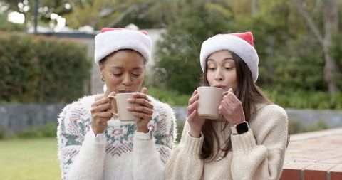 Diverse women wearing santa hats sipping steaming mugs in cozy backyard holiday moment