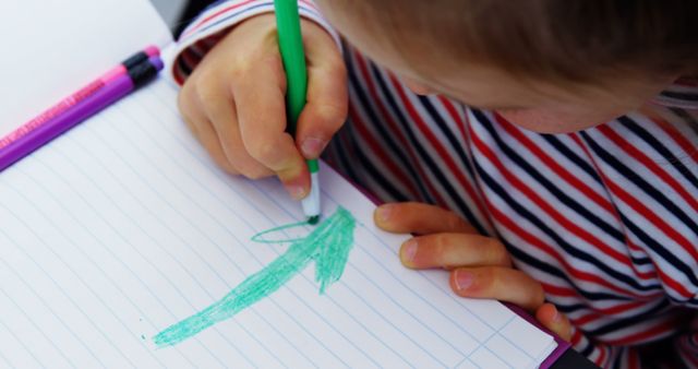 Close-up of a child's hand drawing with a green marker on a lined notebook. The child is wearing a striped shirt, focusing on their creative activity. Useful for educational materials, child development resources, art and craft tutorials, parenting blogs, and school promotions.