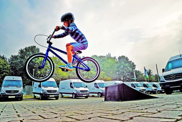 Bright image showing young kid performing a bike stunt in a parking lot with several vans in the background. Perfect for use in advertising outdoor activities, kids' safety gear, biking events or automotive compositions.