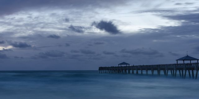 Calm ocean scene with a long pier extending into the water at dusk. Cloudy sky creates a dramatic backdrop, enhancing the tranquil mood. Ideal for use in designs focused on tranquility, relaxation, travel, and nature. Can be used for print and digital media to evoke a peaceful, serene ambiance.