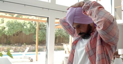 Man adjusting turban indoors in casual plaid shirt
