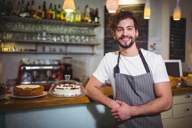 Young male waiter standing at a cafe counter, smiling warmly. He is wearing an apron and has cakes displayed on the counter. Ideal for use in advertisements, hospitality industry promotions, and articles about cafes or customer service.