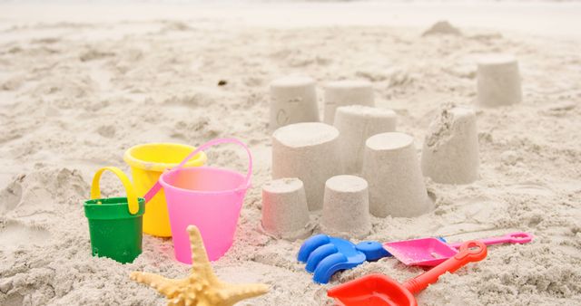 Colorful sand toys such as buckets and shovels are placed next to sand castles on a sunny beach. A starfish lies in the foreground, emphasizing the seaside setting. Ideal for portraying summer vacations, family fun, childhood playtime, and relaxation by the sea.