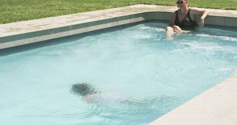 Mother relaxing at pool edge while child practicing swimming in private backyard