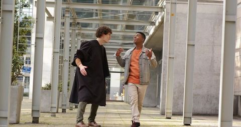 Two students walking and laughing under metal canopy on campus one wearing graduation gown
