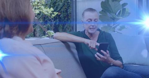 Mature woman teaching friend smartphone use on sofa, casual conversation with blue light flare