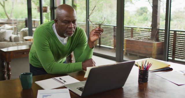 Senior man working from home, using laptop at desk. Wearing green sweater, looking focused. Office supplies and coffee mug on desk. Suitable for topics on remote work, retirement, home office setups, modern technology, and senior lifestyle.