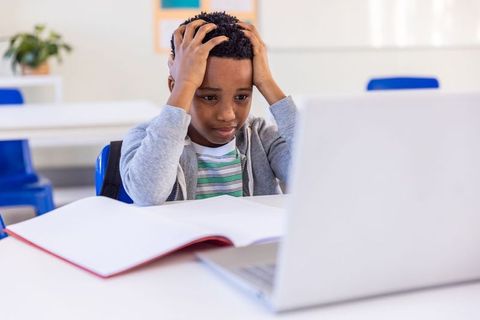 African american boy struggling with online learning holding head at laptop in classroom
